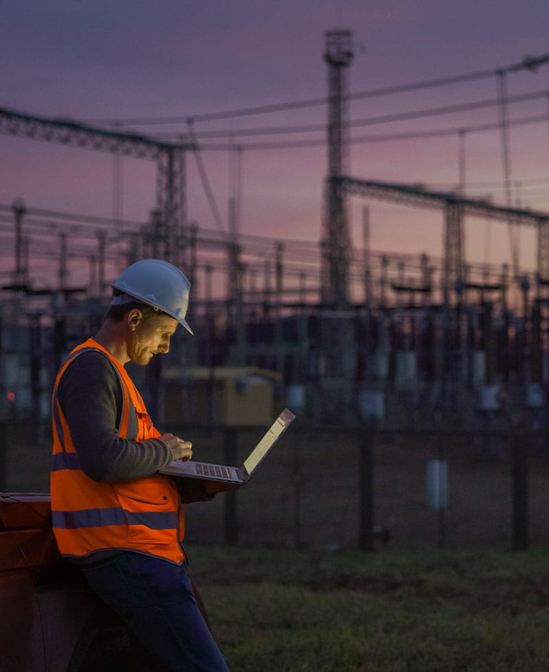Un ingeniero de servicios públicos, con casco y chaleco de seguridad, revisa un ordenador portátil en una subestación eléctrica al atardecer.