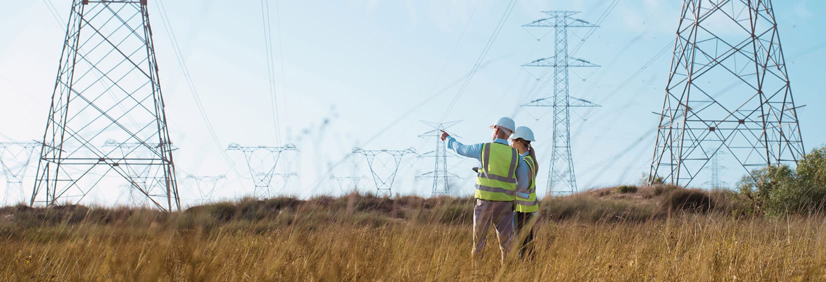 Dos trabajadores de servicios públicos con chalecos y cascos de seguridad inspeccionan torres de transmisión eléctrica en un campo cubierto de hierba.